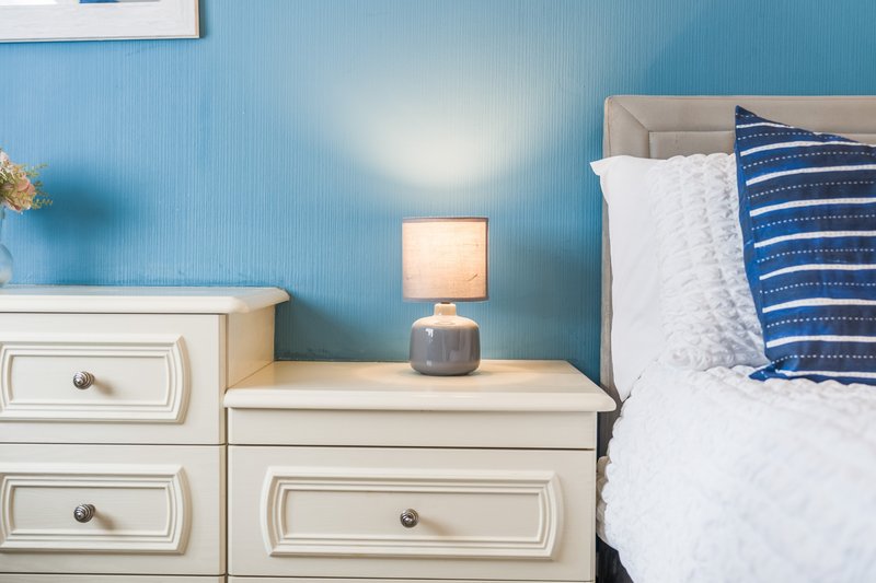 Bedroom with blue textured wall, cream bedside table, small lamp, and striped blue pillow