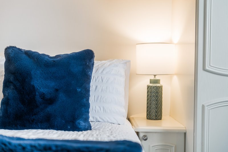 Bedroom corner with blue velvet pillow, white bedding, and table lamp on nightstand