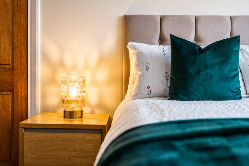 Bedroom with upholstered headboard, teal velvet cushion, white bedding, wooden nightstand with decorative lamp