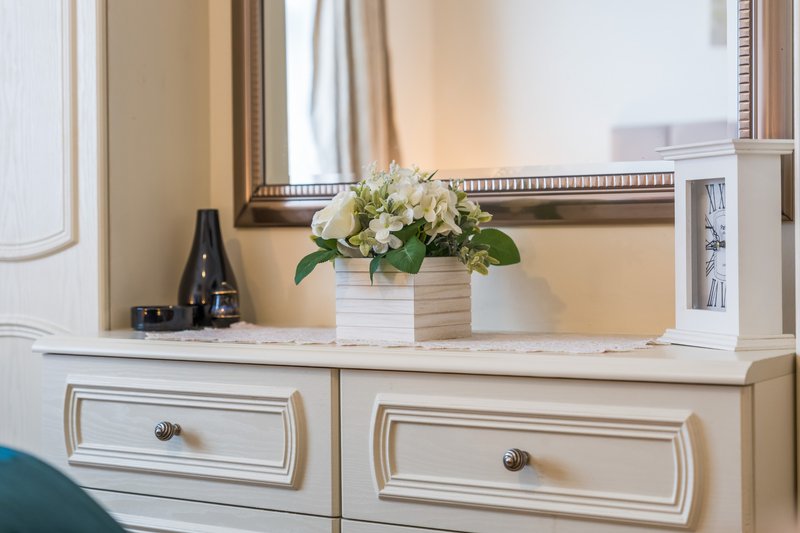 Bedroom dresser with white drawers, mirror, potted flowers, and dark bottle decor