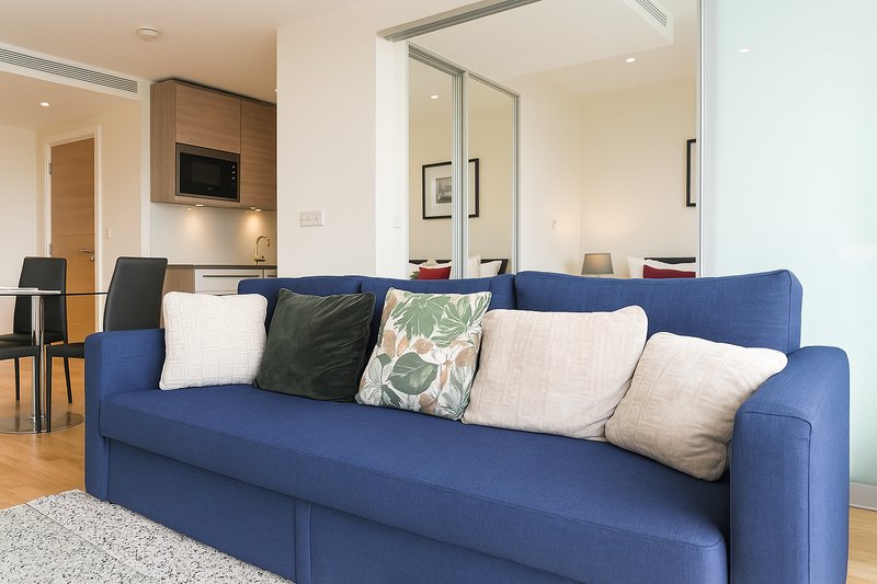 Modern open-plan living room with blue sofa, decorative cushions, and kitchen area visible