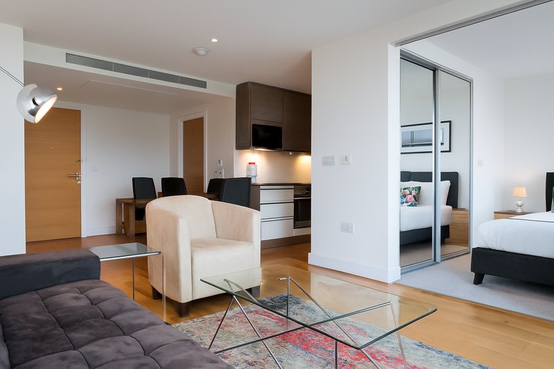 Modern open-plan studio with cream armchair, dark sofa, glass table, and kitchenette visible in background
