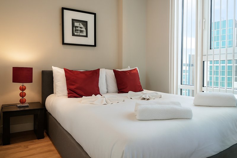 Modern bedroom with double bed, red pillows, white linens, bedside lamp and city view