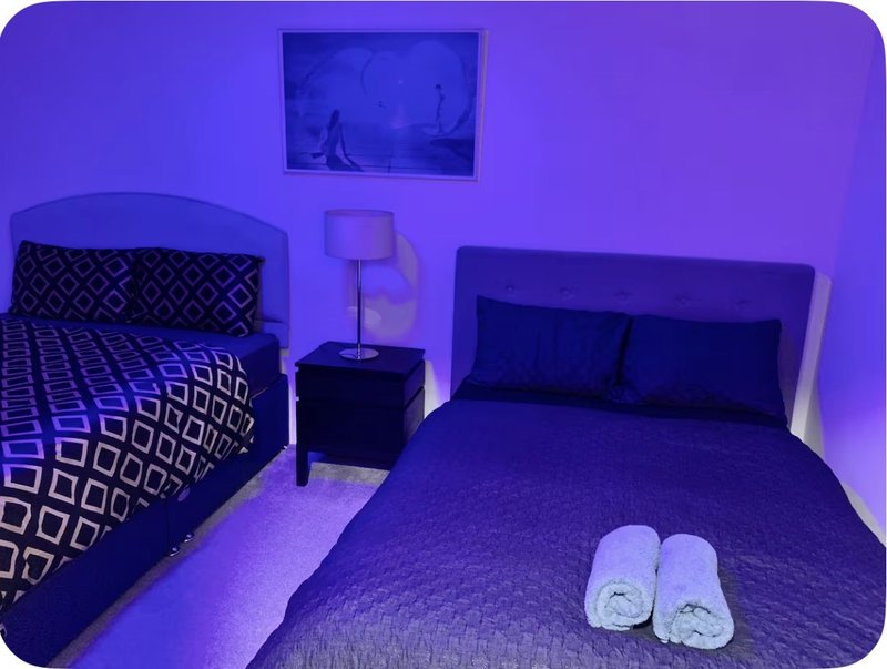Bedroom with two beds, purple ambient lighting, bedside table and wall-mounted artwork