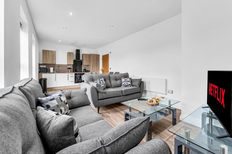 Open-plan living room with grey sofas, glass coffee table, TV, and kitchen area visible in background
