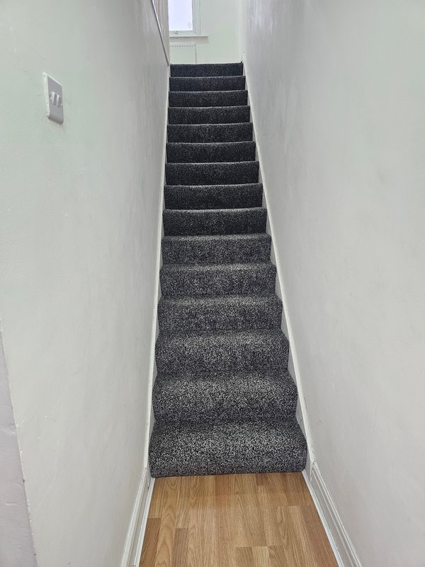 Narrow staircase with dark gray carpet and white walls, wooden floor at base