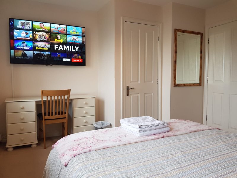 Bedroom with double bed, wooden desk and chair, chest of drawers, wall-mounted TV showing menu interface