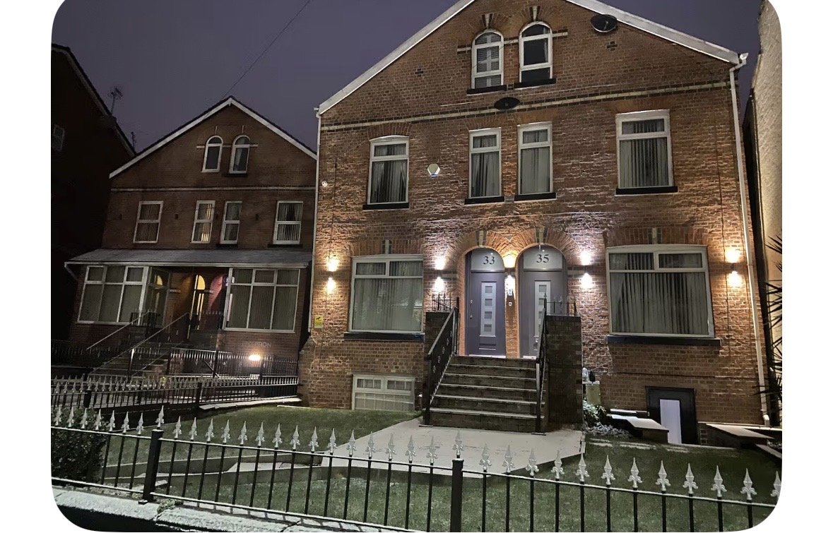 Brick building exterior at night with lit entrance, metal railings and adjacent structures