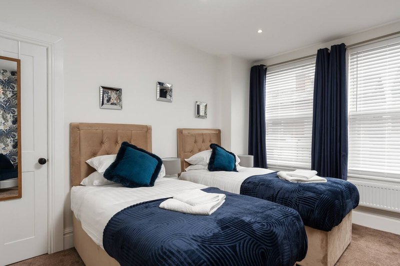 Twin bedroom with two beds, blue bedding, teal cushions, and large windows with dark curtains