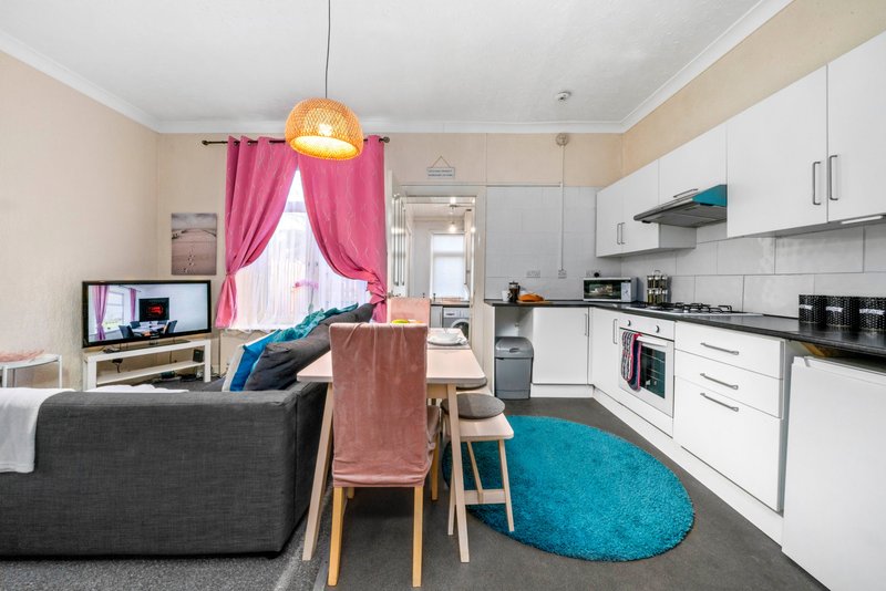 Open-plan kitchen and living area with white cabinetry, dining table, grey sofa and pink curtains
