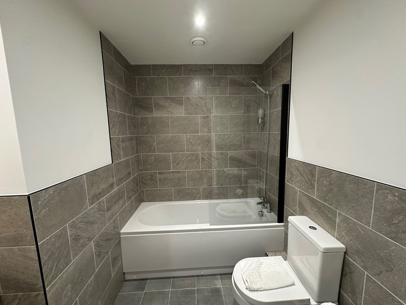 Modern bathroom with gray tile walls, white bathtub, toilet, and recessed ceiling light