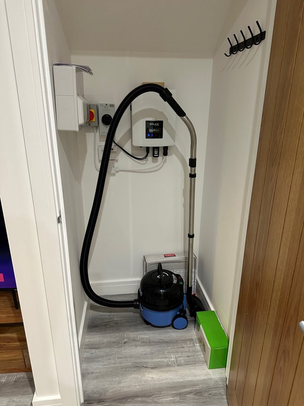 Utility closet with wall-mounted electrical box, black hose, blue and black vacuum cleaner, and green storage box