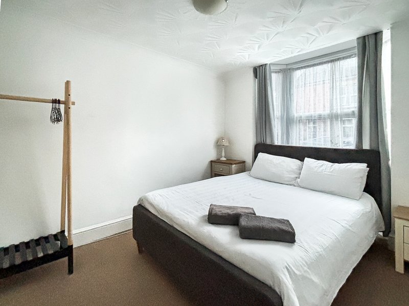 Bedroom with double bed, white bedding, dark headboard, wooden coat stand and window with grey curtains