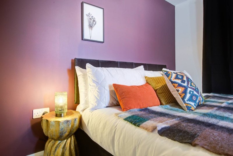 Bedroom with purple walls, double bed, colorful pillows and bedding, wooden nightstand with lamp
