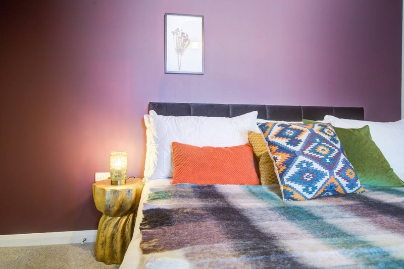 Bedroom with purple walls, double bed, colorful pillows, patterned bedding and bedside lamp