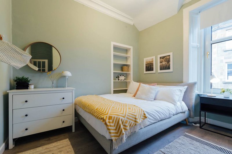 Bright bedroom with double bed, white dresser, round mirror, and yellow throw blanket