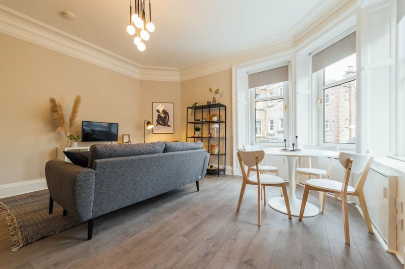 Bright living room with grey sofa, wooden dining chairs, large windows and pendant lighting
