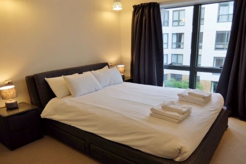Modern bedroom with queen bed, cream bedding, dark headboard, and floor-to-ceiling window with city view
