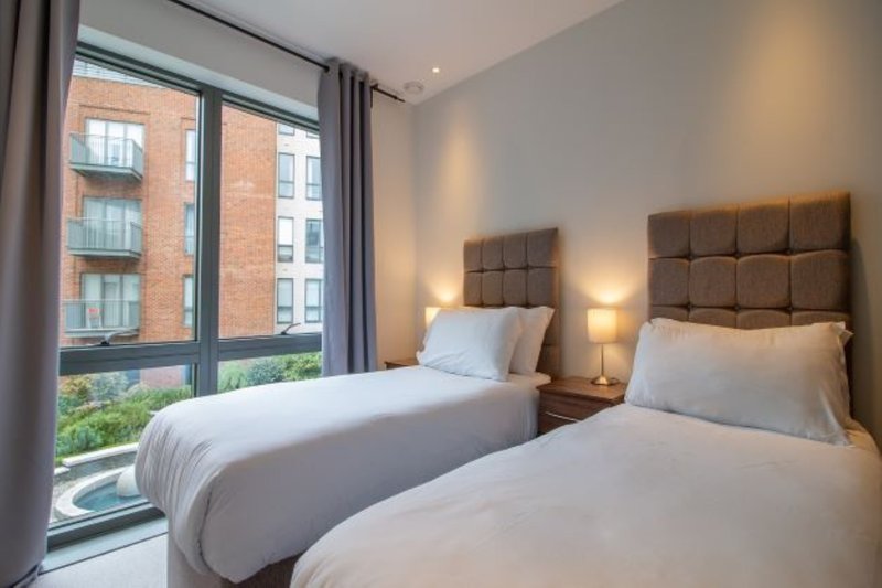 Modern twin bedroom with two single beds, brown headboards, bedside lamps and large window overlooking brick building