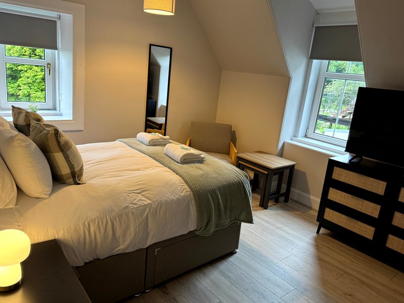 Bright attic bedroom with double bed, cushions, mirror, TV, and garden views from windows