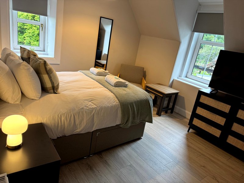 Bright bedroom with double bed, cushions, bedside lamp, mirror, and windows overlooking garden