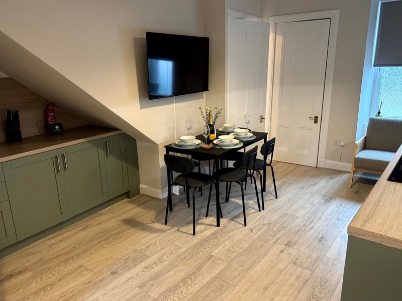 Open-plan kitchen and dining area with sage green cabinetry, black dining table, wall-mounted TV, and wooden flooring