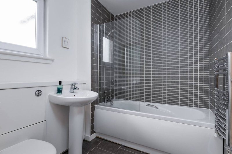 Modern bathroom with pedestal sink, white bathtub, grey tile walls and window