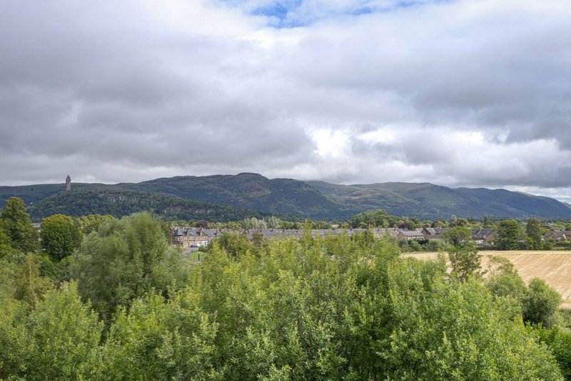 Scenic rural landscape view with tree-lined foreground, distant village, and mountains under cloudy sky