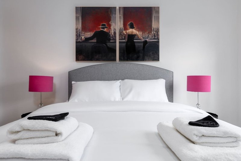 Modern bedroom with gray upholstered headboard, white bedding, pink lamps, and wall art