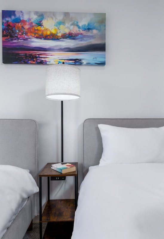 Bedroom with two gray upholstered beds, white bedding, wooden nightstand with lamp, and colorful landscape artwork above