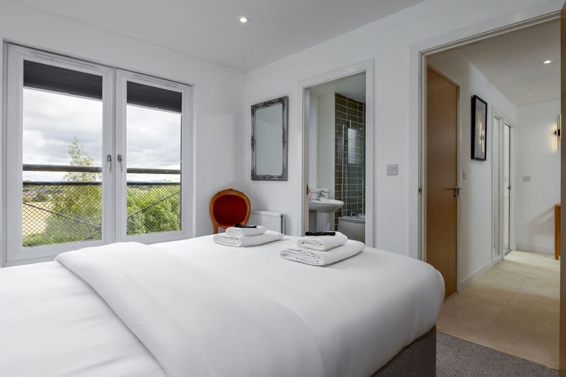 Bright modern bedroom with white bed, large windows overlooking garden, and ensuite bathroom entrance