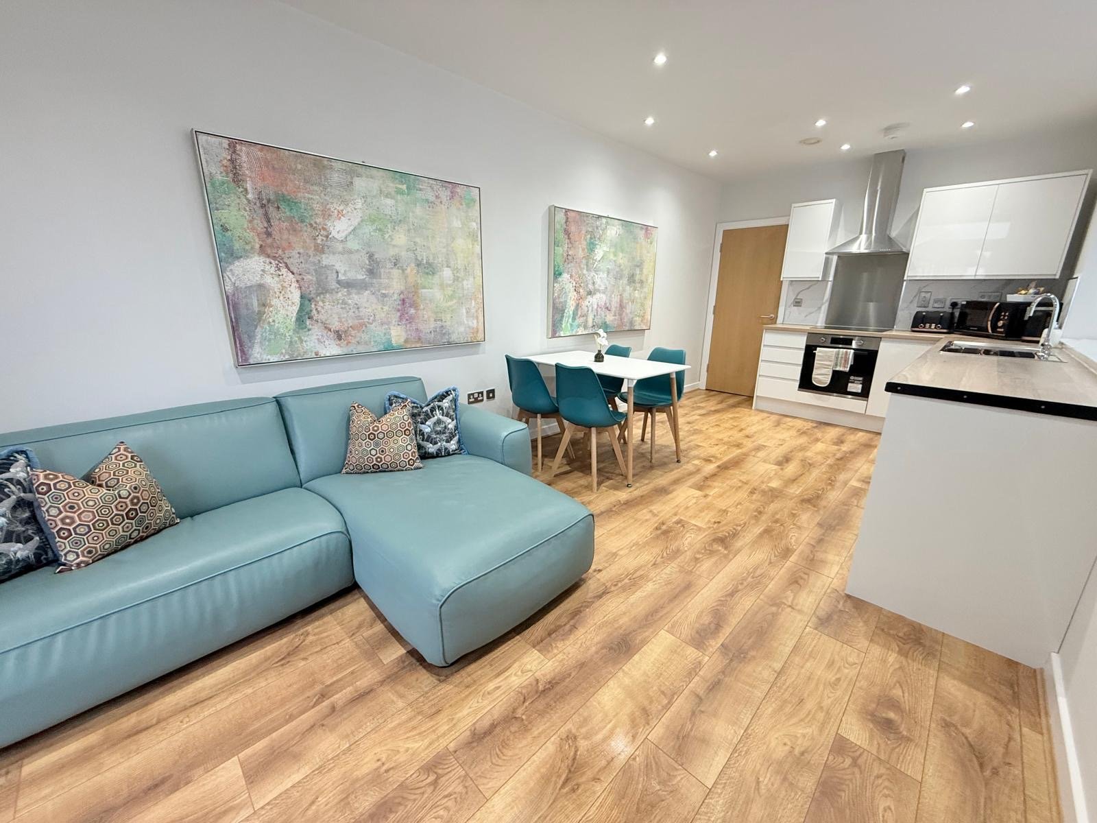 Open-plan living area with turquoise sofa, dining table, and white kitchen with wooden flooring