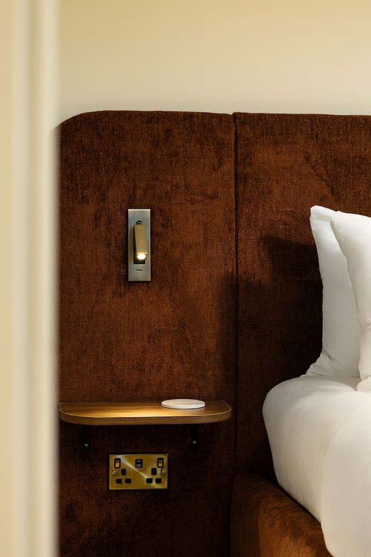 Bedroom detail with brown upholstered headboard, white pillow, bedside shelf with lamp and candle