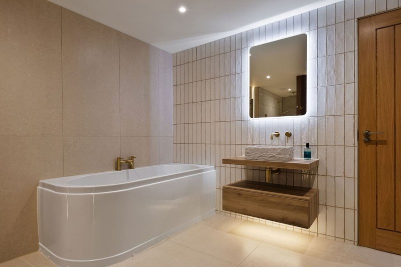 Modern bathroom with curved white bathtub, illuminated mirror, wooden vanity and tiled walls