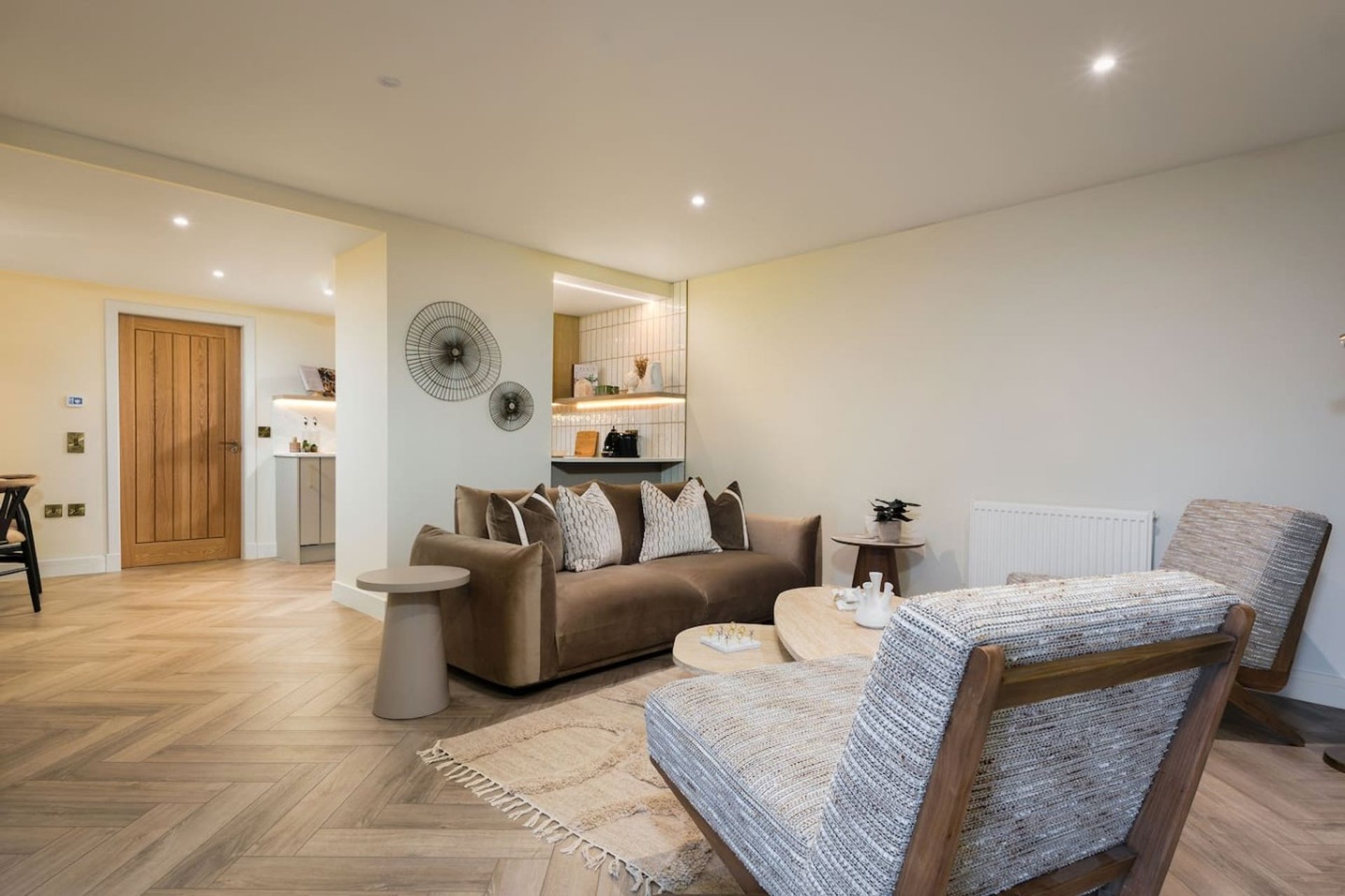 Open-plan living area with brown sofa, upholstered armchair, wooden flooring and kitchenette