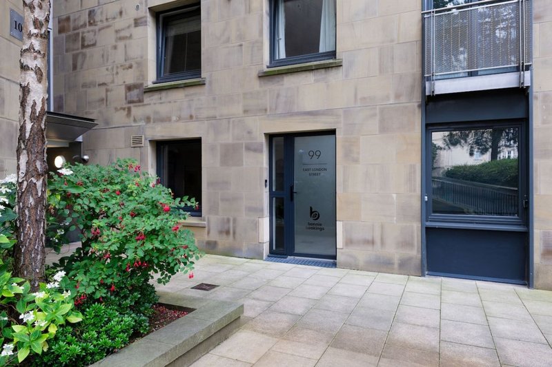 Stone-built apartment building exterior with entrance door, windows, and front courtyard garden