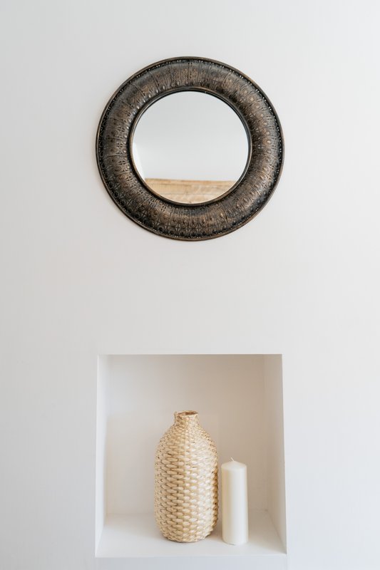 White wall with round textured mirror and built-in niche displaying woven vase and candle