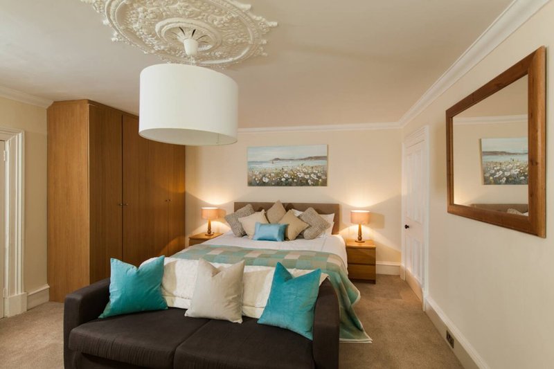 Spacious bedroom with double bed, teal cushions, black sofa, wooden wardrobe and pendant light