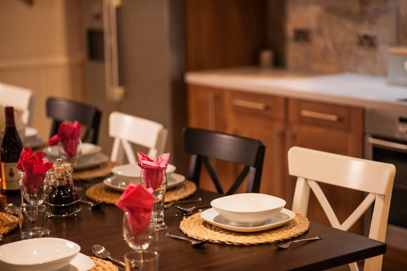 Dining table set for a meal with place settings, red flowers, and kitchen visible in background