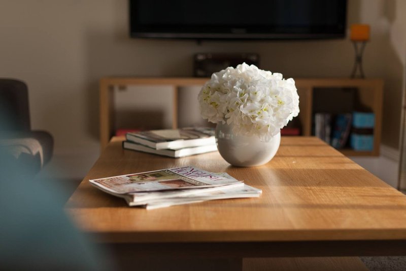 Living room with wooden coffee table, white flower arrangement, magazines, and wall-mounted TV