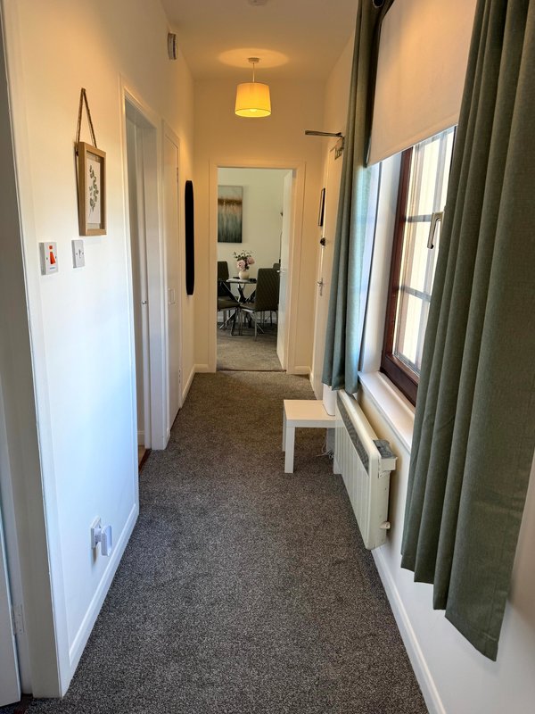 Narrow hallway with grey carpet, white walls, pendant light, and green curtains on window