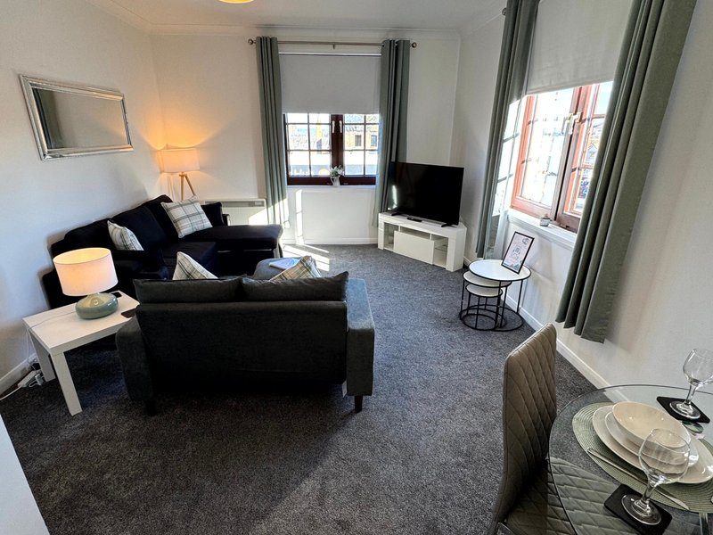 Bright living room with black L-shaped sofa, TV, dining area, and glass doors to balcony