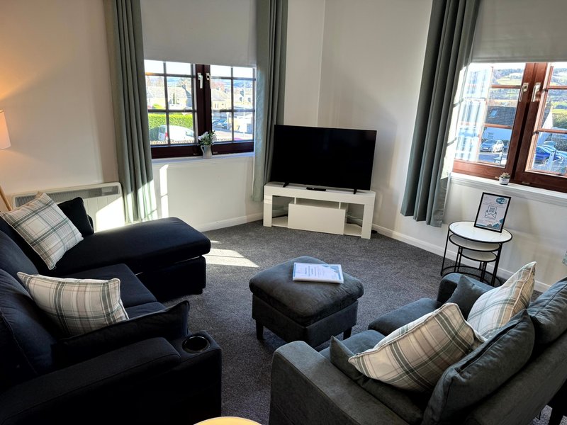 Bright living room with dark sofas, patterned cushions, ottoman, TV and large windows