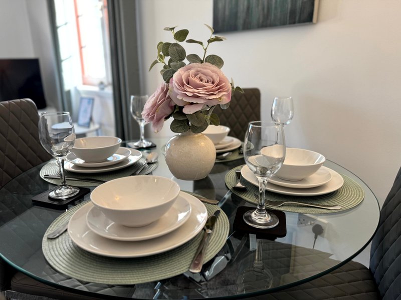 Dining table set for four with white plates, glasses, and pink rose centerpiece