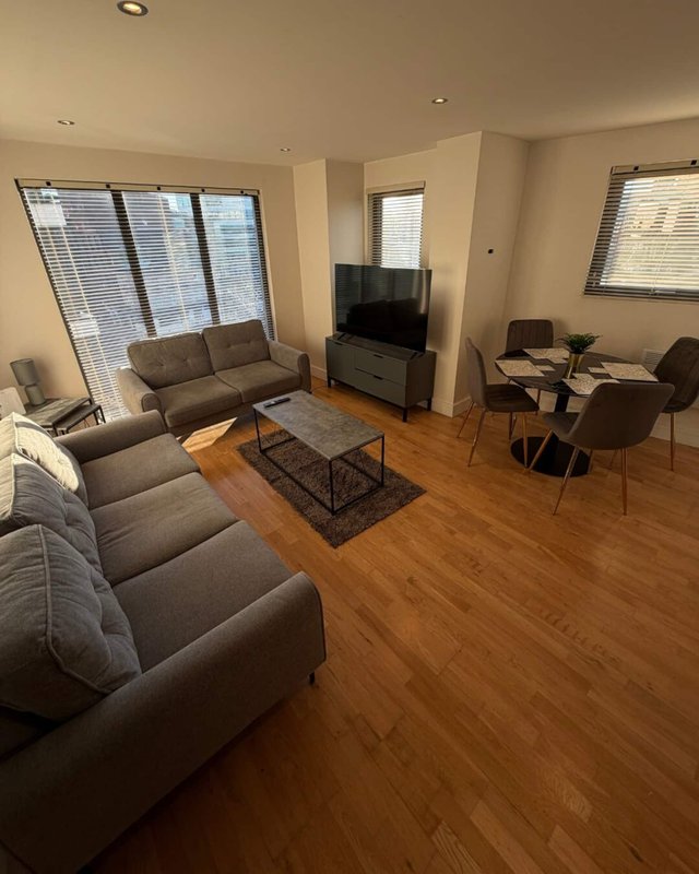 Spacious living room with grey sofas, wooden flooring, TV, and large windows with blinds
