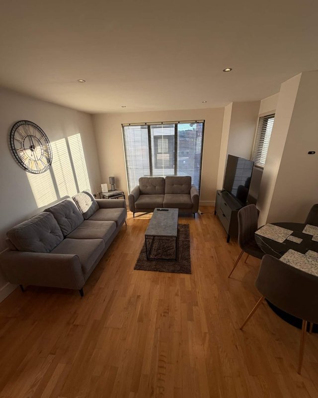 Modern living room with wooden floors, grey sofas, wall clock, and floor-to-ceiling windows