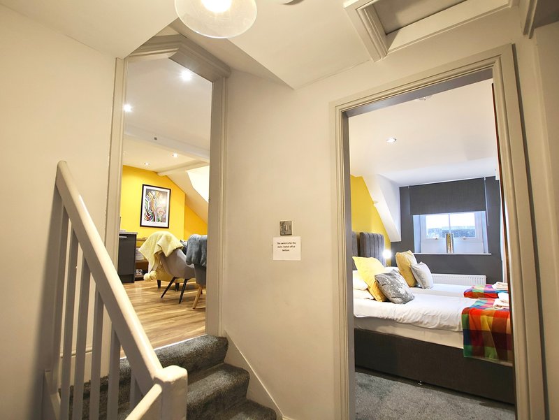 Hallway with staircase leading to yellow-walled living area and bedroom with double bed visible through open door