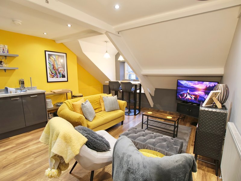 Bright attic living room with yellow accent wall, yellow sofa, grey armchair, TV, and wooden flooring