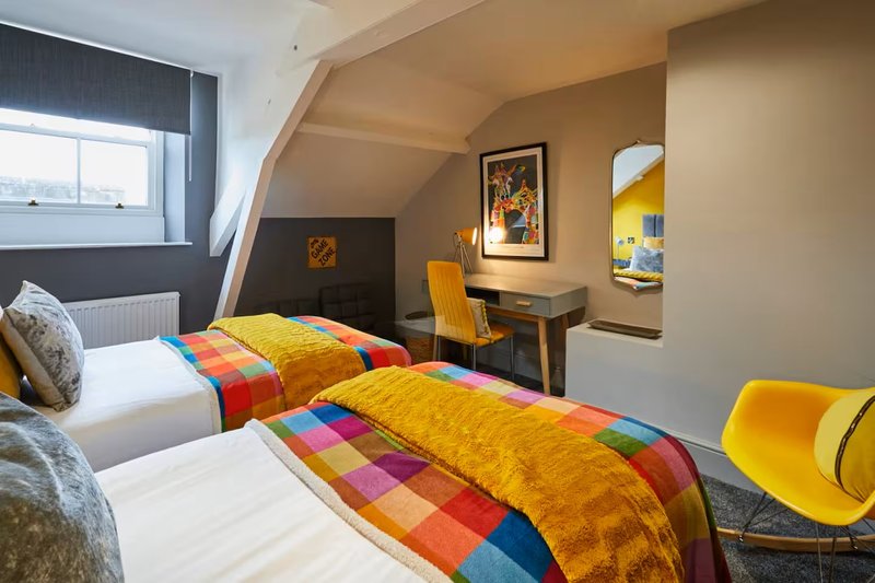 Bright attic bedroom with two beds, colorful patchwork bedding, desk, chair and framed artwork