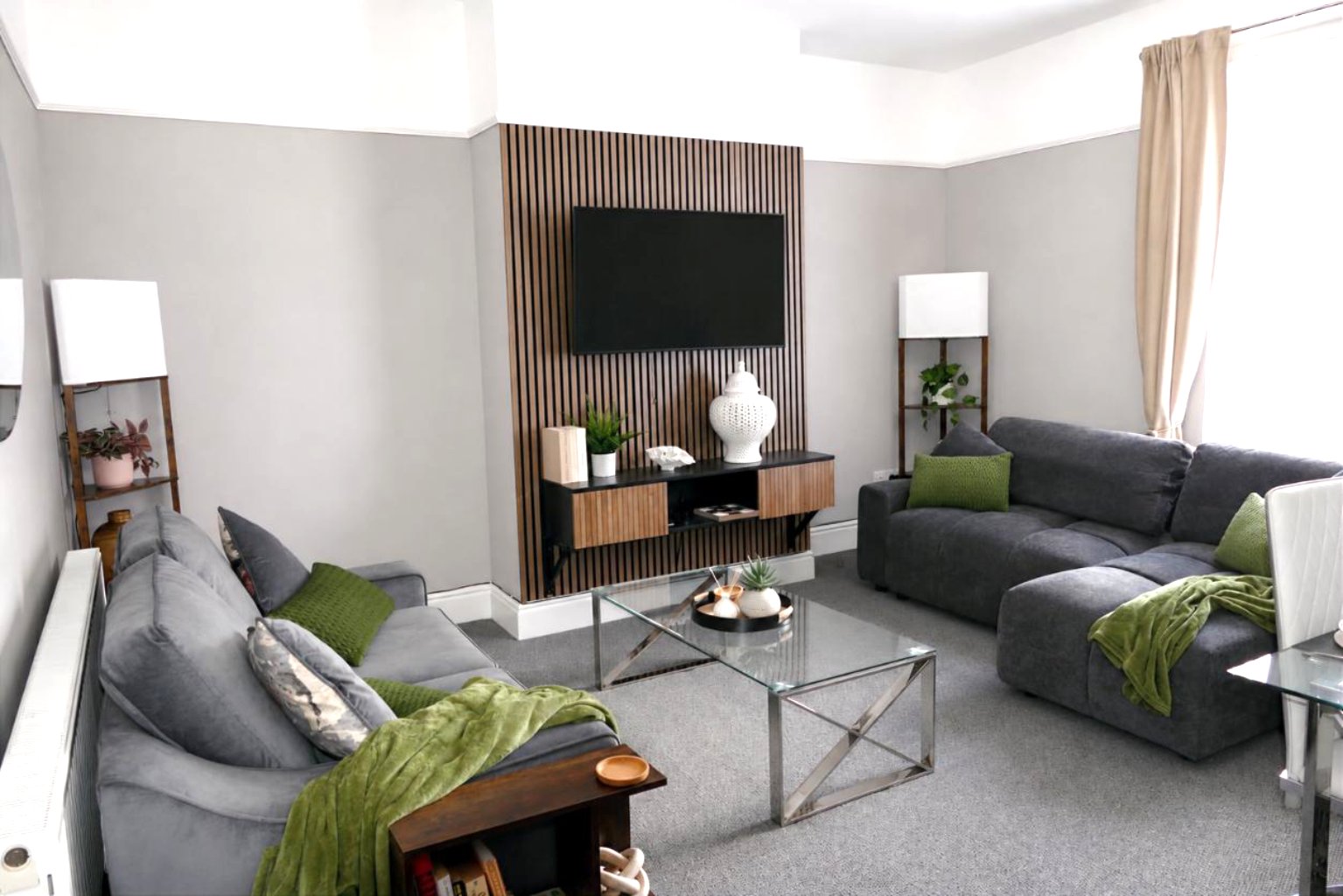 Modern living room with  two gray sofa beds, green throws, wall-mounted TV, wooden console table, and glass coffee table